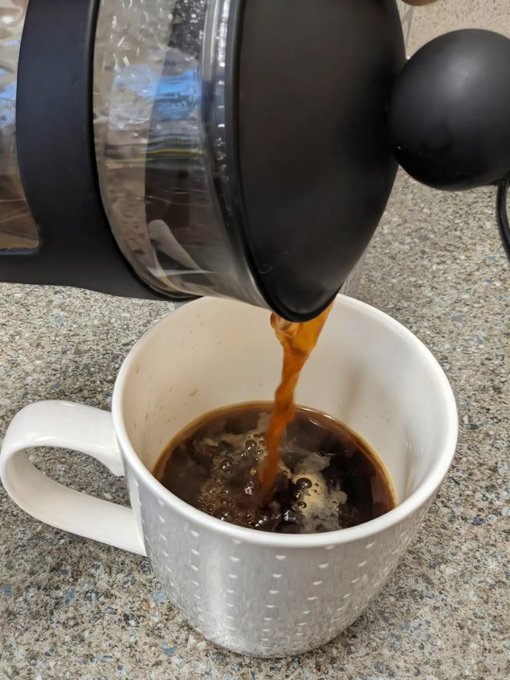 The image shows a white coffee cup filled with hot chocolate, or coffee, being poured into it by an electric coffee maker. The cup has a handle on one side for easy holding. The background of the image is blurred, but there are some words visible that seem to be related to the post "GM future millionaires Who'S busy buying the dip today?".