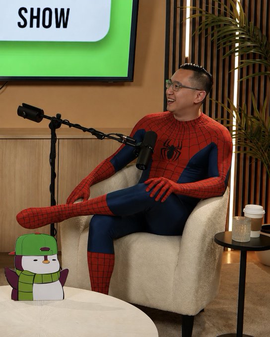 A man dressed in a Spider-Man costume sitting in a beige armchair on a talk show set, smiling next to a microphone, with a 'SHOW' logo visible in the background and a penguin plushie wearing a green hat and scarf in the foreground.