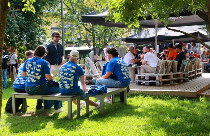A group of people wearing matching blue t-shirts sit on a bench at an outdoor festival, watching an event with trees and other attendees in the background.