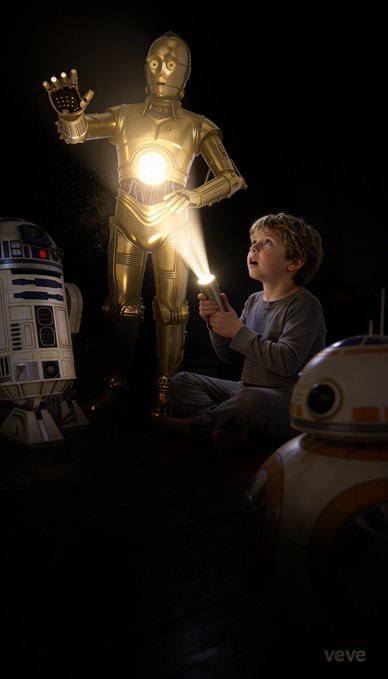 A young boy in pajamas sits on the floor shining a flashlight at a life-size C-3PO figure, with R2-D2 and BB-8 nearby in a dark room.