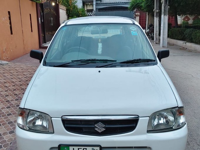A white Suzuki sedan is parked on a residential street, featuring the Suzuki logo on its grille and a partially visible green license plate beginning with "LES." The image captures the pride of the owner celebrating the acquisition of their first car, with a small decorative charm hanging from the rearview mirror.