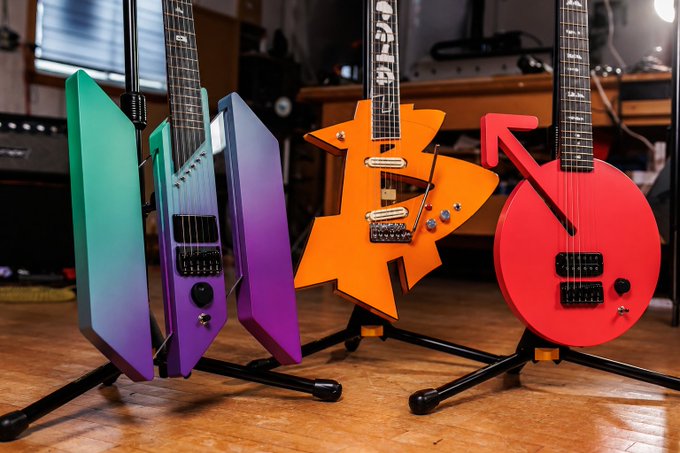 Four uniquely shaped electric guitars in teal, purple, orange, and red stand on display stands in a studio.