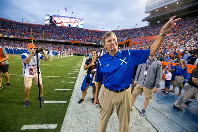 A man in a blue polo shirt and khaki pants waves to a crowd while walking on the sidelines of a football stadium field, surrounded by fans and media.