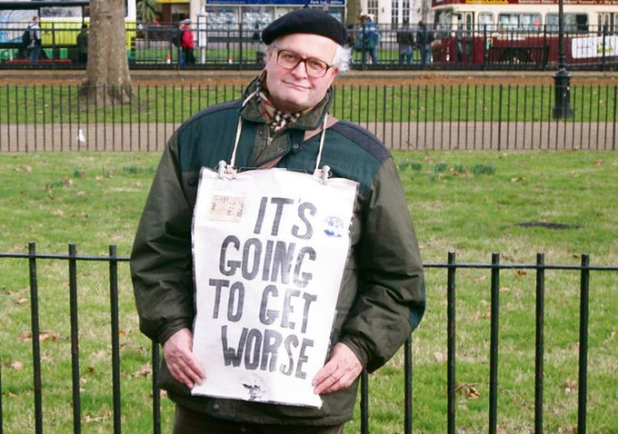 An older man wearing a beret and glasses stands in a park holding a large sign that reads "IT'S GOING TO GET WORSE."