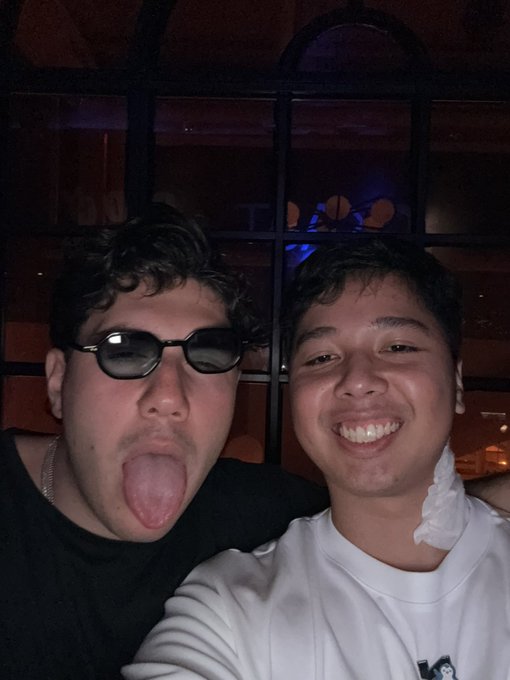 A selfie of two young men smiling in a dimly lit venue, one wearing sunglasses and sticking out his tongue, the other in a white hoodie.