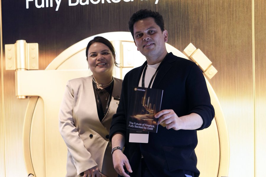 A man and woman posing together in front of a large gold vault backdrop that reads 'Fully Backed By Gold', with the man holding a GoldCava booklet titled 'The Future of Finance. Fully Backed by Gold'.