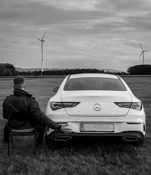 A black-and-white photograph of a man sitting on a chair facing away, his hand resting on the rear of a white Mercedes-Benz sedan parked in a field with wind turbines in the background.