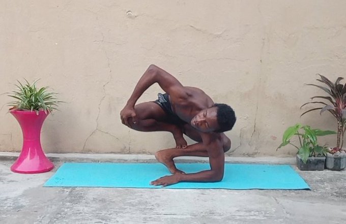 A shirtless man in dark shorts performing an advanced contortion or acrobatic pose on a blue yoga mat, with plants on either side.