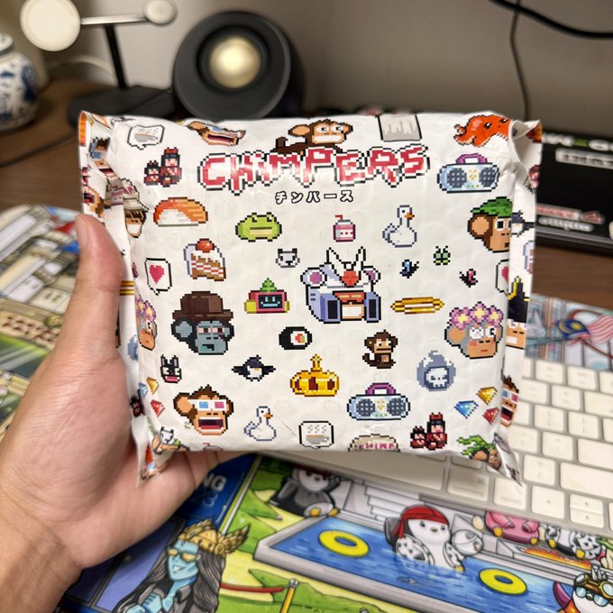A hand holds a package labeled "CHIMPERS" with Japanese text (チンパーズ), featuring a colorful pattern of pixel-art monkey characters and various items, displayed over a matching illustrated desk mat.