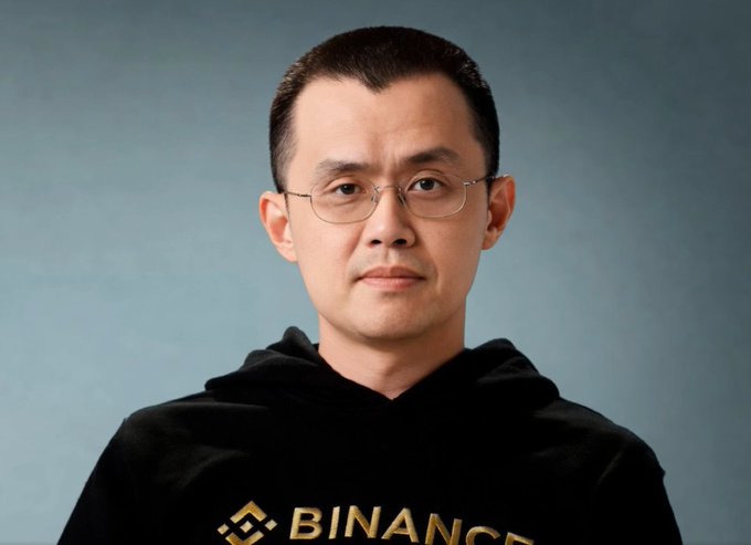 The image features a man wearing glasses, a hoodie with "BINANCE" written on it, and a gold logo. The man appears to be looking directly at the camera while posing for the photo. The background of the image is a solid blue color that contrasts with the man's attire. There are no other objects or texts in the image besides the man and his clothing.
