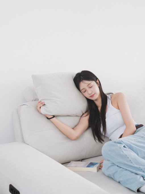 A young woman with long dark hair resting her head on a light gray couch pillow, wearing a white tank top and loose jeans, with an open book beside her on the couch in a minimalist white room.
