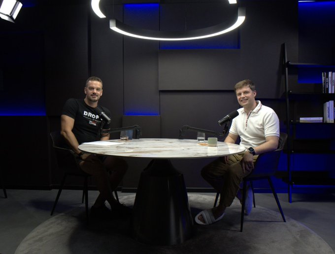 Two men are seated at a round marble table in a sleek studio with blue-lit walls and a distinctive circular overhead light, with one wearing a "DROP" branded t-shirt and both positioned behind microphones for a podcast recording. This image illustrates the post's announcement of a DROPS podcast episode where founder Michael H. sits down with Kevin to discuss the candid, unpolished story behind the launch.