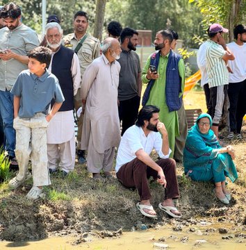 The image shows a group of people standing around, with some sitting down near a body of water. There are at least 12 individuals visible in the scene, including both men and women. The majority of them appear to be engaged in conversation or observing something happening nearby. One person is holding a cell phone, possibly capturing a moment or checking for updates on social media.

The setting seems to be outdoors, with people gathered around a pond or riverbank. There are also some objects present in the scene, such as backpacks and handbags placed near the group of people. The overall atmosphere appears to be casual and relaxed, with everyone enjoying each other's company while taking in their surroundings.
