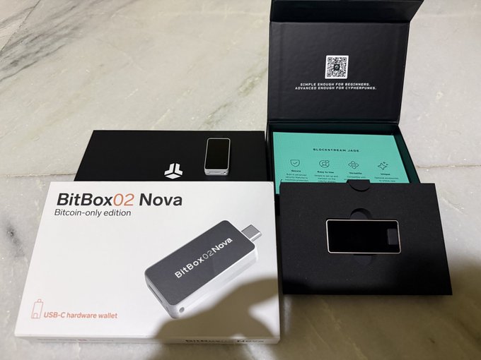 A BitBox02 Nova Bitcoin-only edition hardware wallet is displayed alongside a Trezor-style device in its open black packaging, with the teal informational card visible. The box clearly reads "BitBox02 Nova" and "USB-C hardware wallet," matching the post about the YouTube video featuring both wallets.