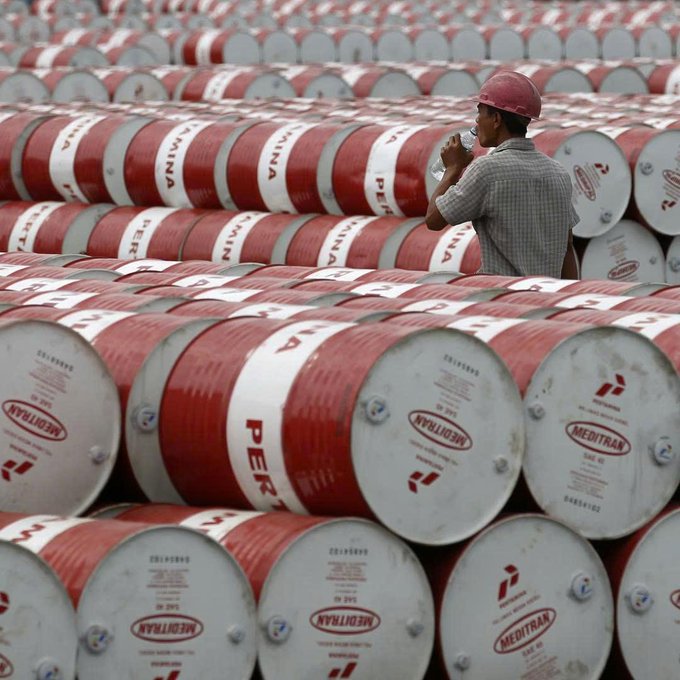 A worker in a red hard hat drinks from a water bottle while standing among rows of red and white oil barrels labeled "MEDITRAN" and "PER", visually representing the Venezuelan oil shipment at the center of the Orlen-USDT payment controversy.