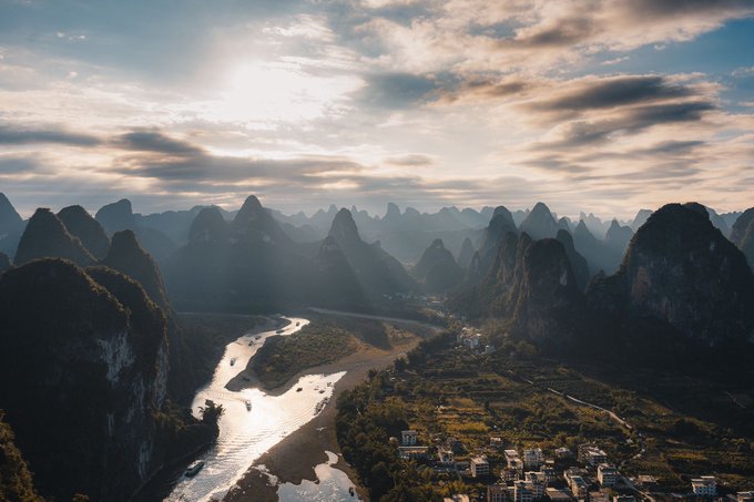 A scenic aerial view of a winding river flowing through misty karst mountain peaks at sunset, with a small village nestled at the base of the hills.