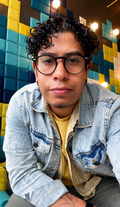 A portrait of a young man with curly hair and round black glasses wearing a light blue denim jacket over a yellow shirt.