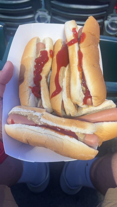 A hand holds a paper tray containing four hot dogs topped with ketchup and mustard, taken from a first-person perspective with the person's legs visible.