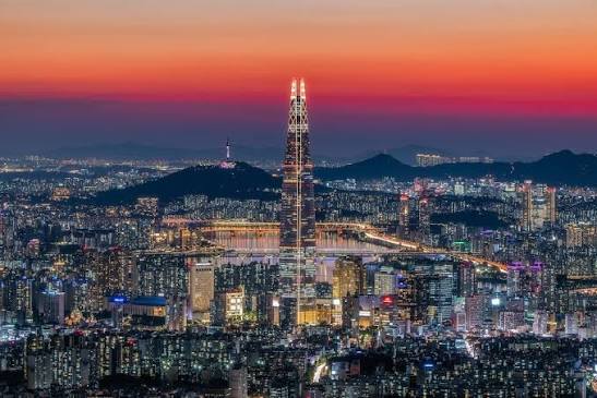 The image shows a city skyline at night, with the Seoul Tower being the tallest structure visible. The tower's lights are shining brightly against the dark sky, creating a stunning contrast. In the background, there are other buildings of varying heights, contributing to the overall urban landscape. The photo is taken from an elevated position, providing a bird's eye view of the city at night.