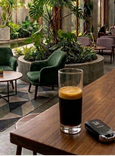 A glass of espresso sits on a wooden table with a Toyota car key fob beside it, with a green armchair and indoor garden visible in the background.
