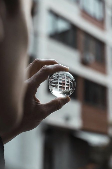 A hand holds a clear crystal ball against a blurred urban backdrop, with a building and trees captured in the sphere's reflection.