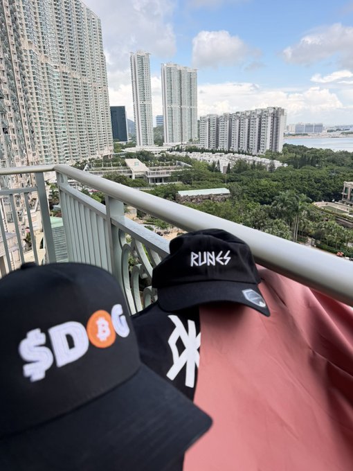 Two black baseball caps (one with 'SD☀G' and another with 'RUN✦S') sitting on a balcony railing with a view of tall residential skyscrapers, greenery, and a cityscape in the background.