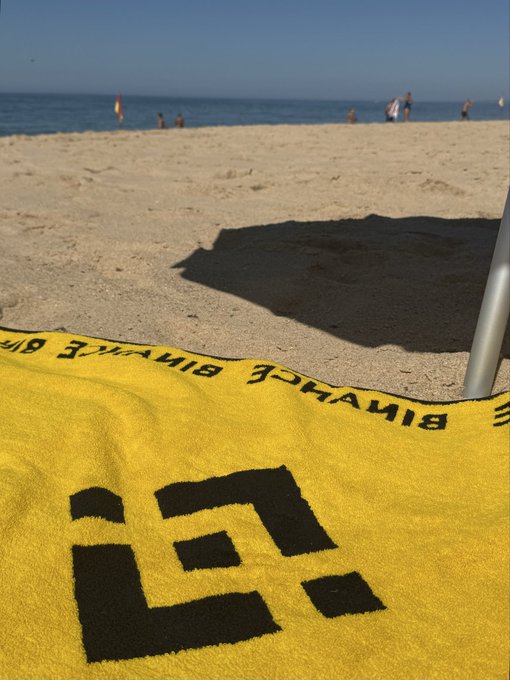 A yellow 'Binance' towel is spread out on a sandy beach with the ocean visible in the background.