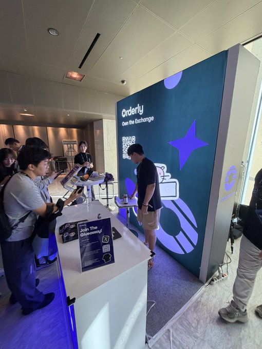 People gathered at what appears to be a trade show or conference booth displaying a large 'Orderly' sign with the tagline 'Own The Exchange' and promotional materials.