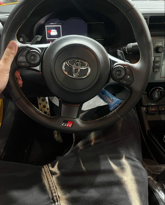 A hand grips a black leather Toyota GR steering wheel with contrast stitching, featuring the "GR" badge at the bottom and the Toyota logo in the center, inside what appears to be a sporty vehicle. The caption "Gn bros 🤍" expresses a friendly goodnight greeting, fitting the casual, selfie-style shot of the driver's seat that feels like a personal sign-off to followers.
