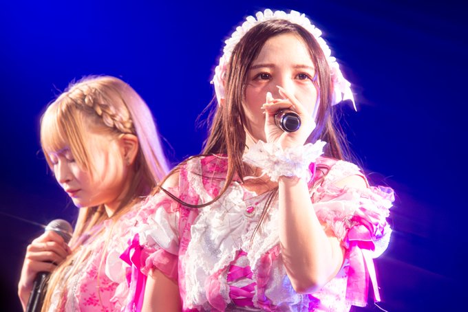 Two young women perform on stage; the one in front sings into a microphone wearing an ornate pink and white ruffled outfit with a floral headband.