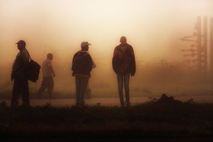 The image shows a group of people walking down a road, with one person wearing a backpack. The scene appears to be foggy or misty, which adds an atmospheric quality to the photo. There are at least four individuals visible in the image, each carrying their own belongings such as backpacks and handbags. The overall mood of the photograph is somewhat mysterious due to the fog and the presence of multiple people walking together.