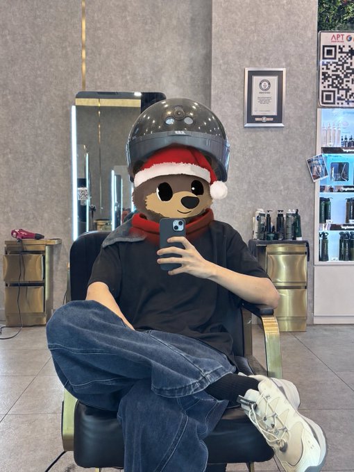 A person taking a mirror selfie wearing a motorcycle helmet with a Santa hat filter, a red scarf, and a bear emoji covering their face, while sitting in a salon chair near a QR code display showing "APT" and an IP reminder sign. The image relates to the post by representing the anonymous social media creator discussing their modest monthly earnings from X (Twitter) creator monetization.