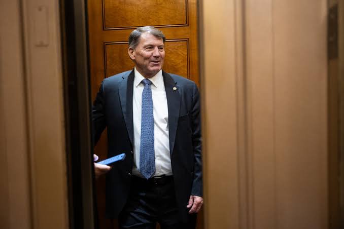 A middle-aged man in a dark suit and blue tie steps through a doorway holding a blue folder, appearing to smile. The image's formal attire and governmental-style hallway setting visually complement the post's news about the new Zcash lobbying organization PGPZ registering in Washington to engage in policy advocacy.