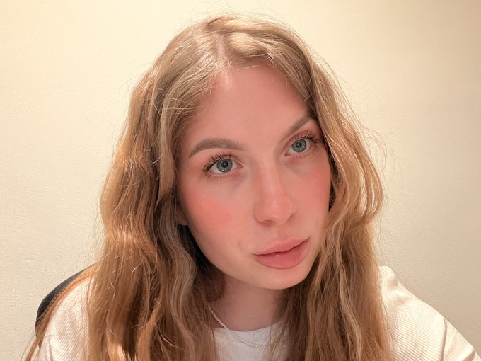 A close-up portrait of a young woman with long wavy blonde hair and blue eyes, wearing a light-colored top against a plain cream background.