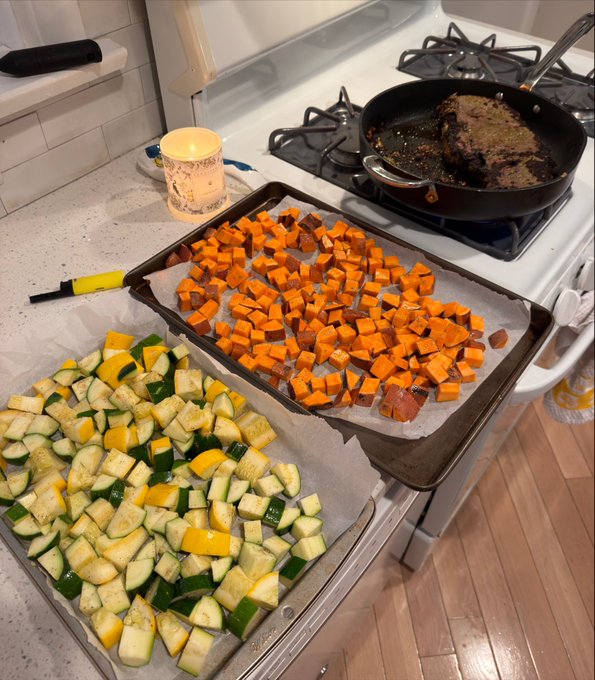 A casual photograph of a kitchen counter and stovetop showing two sheet pans lined with parchment paper; one holds chopped zucchini and yellow squash, while the other contains diced sweet potatoes, next to a frying pan and a mug.