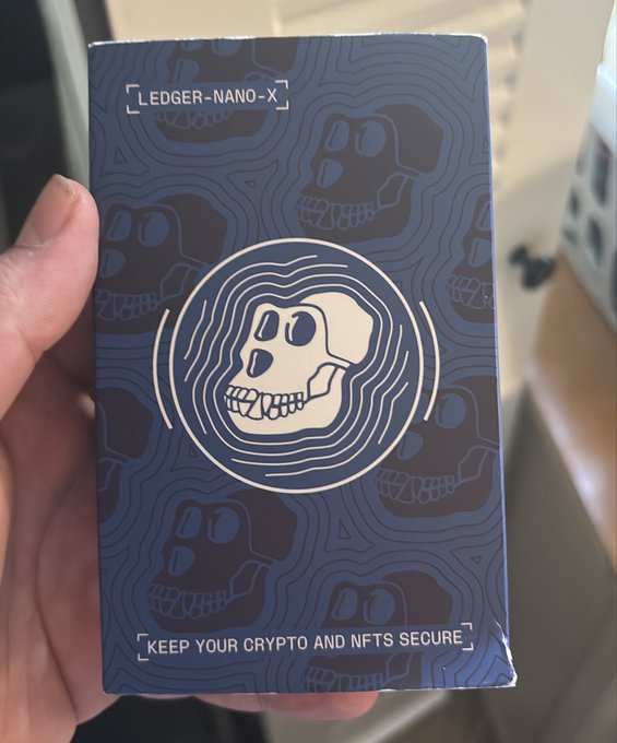 A hand holds up a sealed Ledger Nano X crypto hardware wallet box featuring its signature blue skull design, with text reading "LEDGER-NANO-X" and "KEEP YOUR CRYPTO AND NFTS SECURE." The image fits the "Grail" caption perfectly, as a Ledger is considered the gold standard ("holy grail") for securely storing cryptocurrency and NFTs offline.