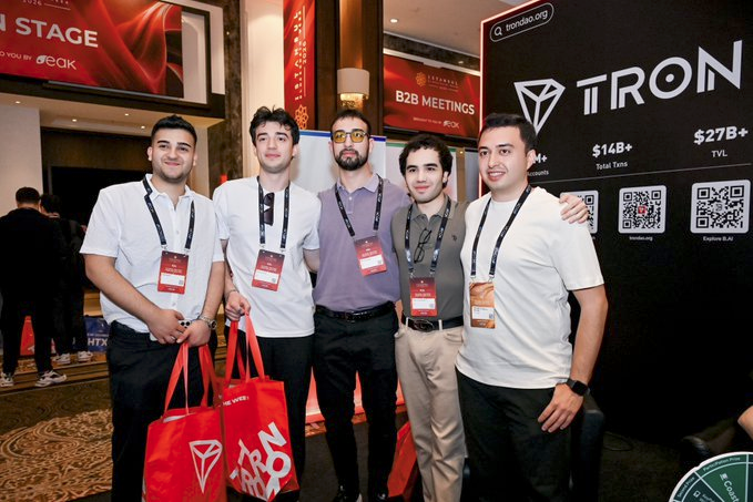 The image features five men standing next to each other with their bags, posing for a photo at an event. They are all wearing name tags that say "TRON" or "IBW", indicating they might be attending a convention related to the tech company or a similar gathering. The group is positioned in front of a booth labeled "TRON" and another one labeled "IBW". The background includes a stage, which suggests this event may have some form of presentation or demonstration taking place.