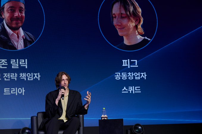 A man with curly hair sits on stage holding a microphone during a panel discussion, with a blue screen behind him displaying portraits and Korean text including '전략 책임자', '공동창업자', '트리야', and '스크드'.