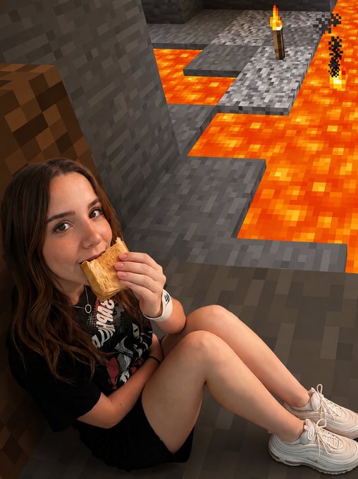 A young woman sits on the floor eating a snack while leaning against a Minecraft-themed wall decorated with pixelated stone blocks and lava.