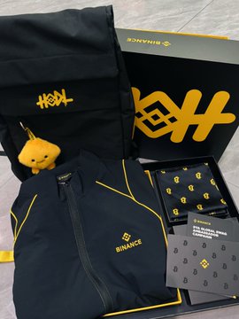 Binance merchandise haul displayed in a branded box, including black and yellow apparel, a small yellow plush bear, socks, and Binance-branded cards.