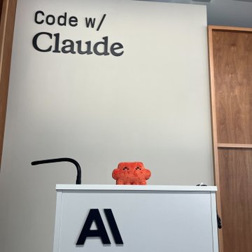 The image shows an Anthropic "Code w/ Claude" event booth, featuring the orange Clawd mascot plush toy alongside the bold "AI" branding on a podium, tying into the $CLAWD token promotion that references both Anthropic and the Clawd character.