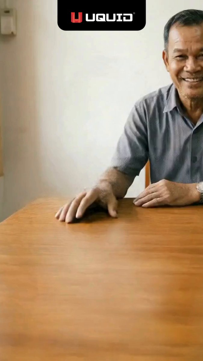 The image features a man sitting at a wooden dining table, smiling for the camera. The background of the photo shows a white wall with a light switch mounted above the table. There are no other people or objects visible in the image, but there is text that reads "A better business can start with a simple integration. UQUID’s API unlocks 600K+ digital products, global stablecoin payments, and instant fulfillment, helping SMEs scale faster with less overhead". The man's smile suggests he might be discussing or sharing this information about the benefits of integrating an API for their business.