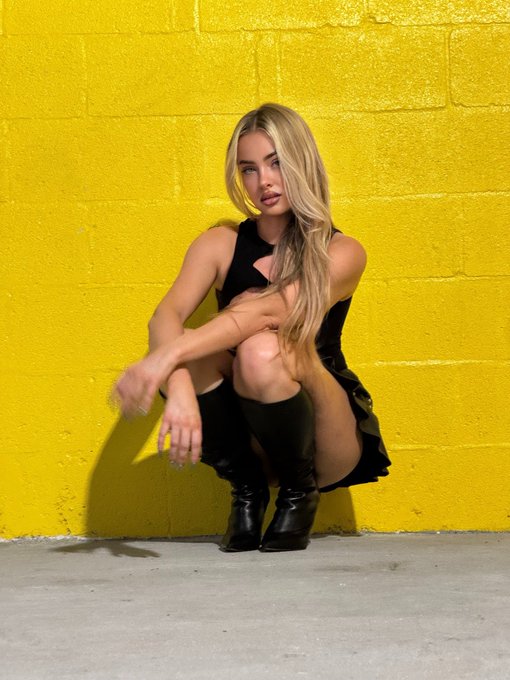 A blonde woman in a black cutout dress and tall boots crouches against a vibrant yellow brick wall, posing confidently for the camera. The bold, sunlit backdrop and stylish pose pair perfectly with a cheerful "gm" (good morning) greeting, exuding a high-energy, fashionable vibe to match the playful post.