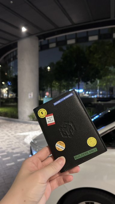 A hand holds a black passport with various stickers (including one for Maersk) up at night in a parking garage or urban setting, with a luxury car partially visible in the background.