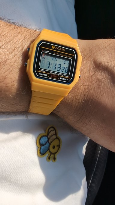 Close-up photo of a person's wrist wearing a yellow ZCASH-branded digital watch displaying 'PM 3:13 21' and 'MO 13 22', with a bee patch visible on a white shirt.