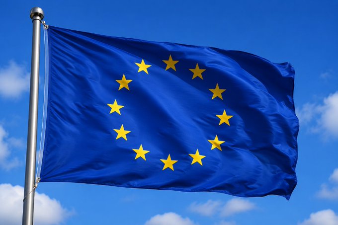 A photograph of the European Union flag with its circle of twelve gold stars on a blue background, waving against a partly cloudy sky.