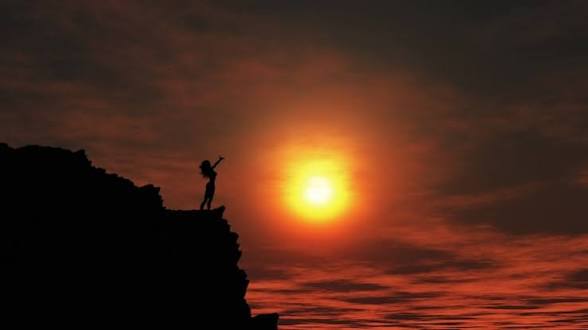 A silhouetted figure stands on a rocky cliff edge with one arm raised, set against a dramatic sunset over the ocean.