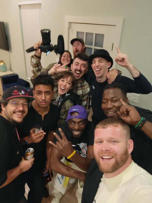 A group selfie of about nine young men in a dimly lit room, smiling and posing enthusiastically, with one holding a camera and another holding a beverage can.