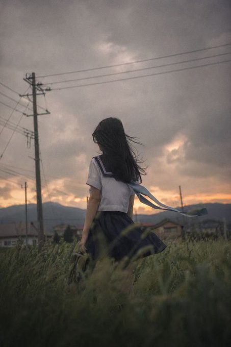 A young woman in a school uniform stands in a grassy field at dusk, her long dark hair blowing in the wind beneath a cloudy sky with power lines stretching overhead. The image's contemplative, transitional mood—capturing a moment of quiet anticipation at twilight—mirrors the post's message about Kaito's return from perceived silence, suggesting a turning point where something dormant comes back into the light.