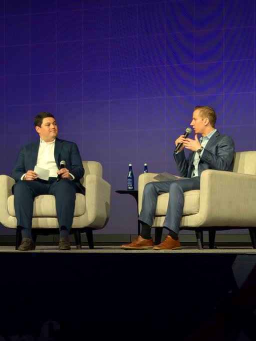 The image shows two men sitting in chairs, one wearing a suit and the other dressed in a t-shirt. They are engaged in conversation with microphones in front of them, suggesting they might be participating in an event or panel discussion. The backdrop features a blue wall with white text that reads "CROPS makes them so bullish on @ethconf". This implies that the men could be discussing the cryptocurrency market and its potential impact on various industries.